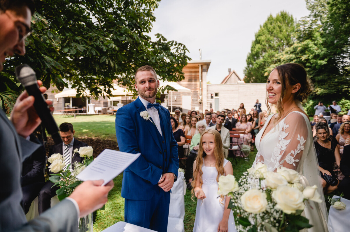 Braut und Bräutigam stehen mit einem jungen Mädchen während einer Hochzeitszeremonie im Freien. Eine Person links spricht in ein Mikrofon, während im Hintergrund Gäste sitzen.