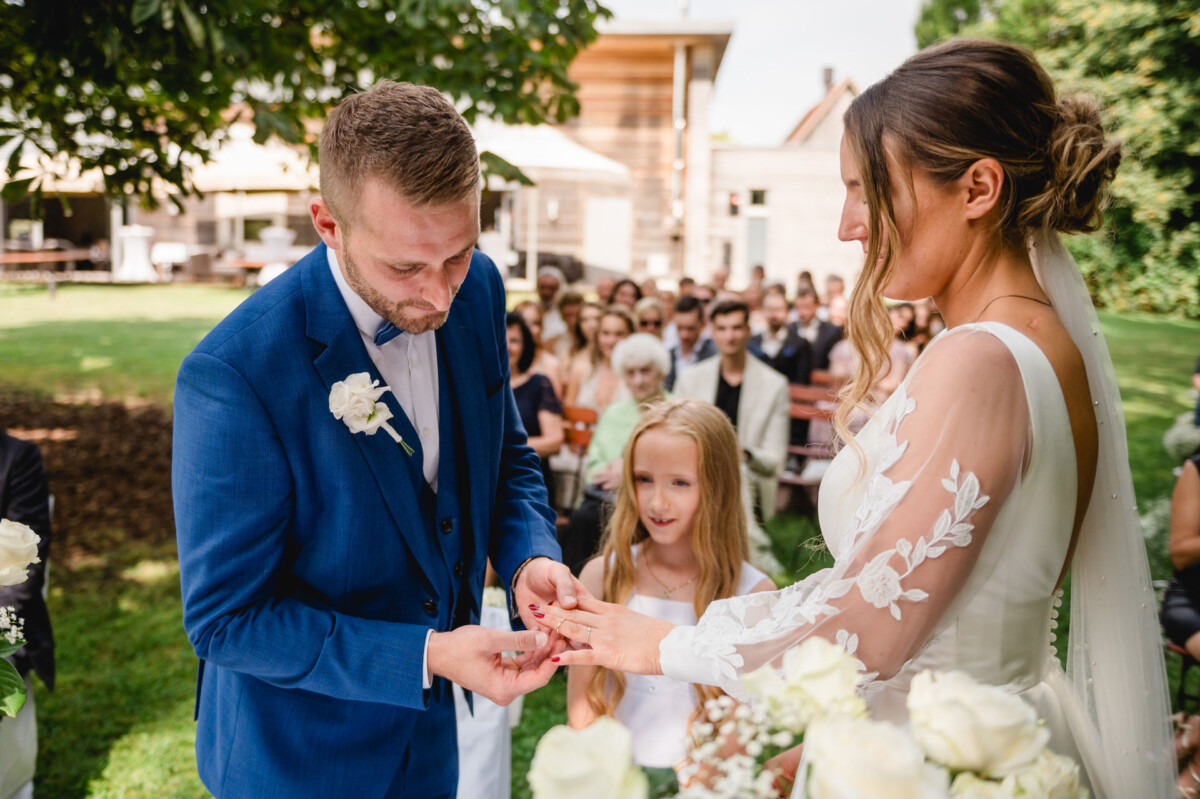 Ein Bräutigam im blauen Anzug steckt der Braut während einer Hochzeitszeremonie im Freien einen Ring an den Finger. Ein junges Mädchen steht daneben.