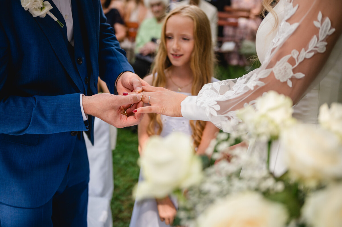 Eine Person im blauen Anzug steckt während einer Hochzeitszeremonie einer anderen Person einen Ring an den Finger. Im Hintergrund beobachtet ein junges Mädchen, umgeben von weißen Blumen, die Szene.