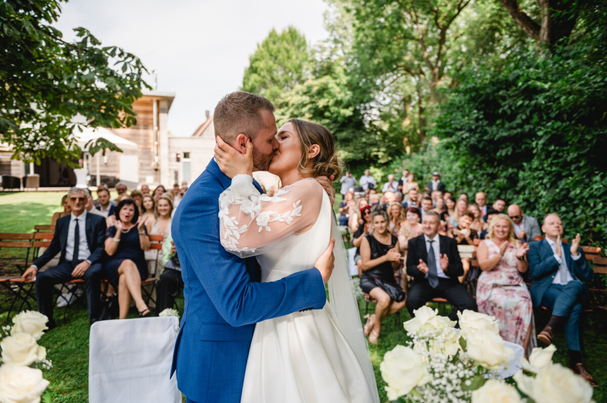 Ein Paar im Hochzeitskleid küsst sich im Freien vor einer sitzenden Menschenmenge, umgeben von Grün und Blumen.