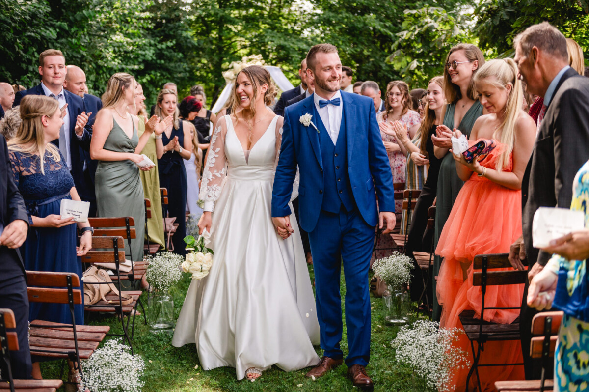 Braut und Bräutigam gehen bei einer Hochzeit im Freien Händchen haltend den Gang entlang. Umgeben von grünem Laub und Holzstühlen stehen die Gäste auf und applaudieren.