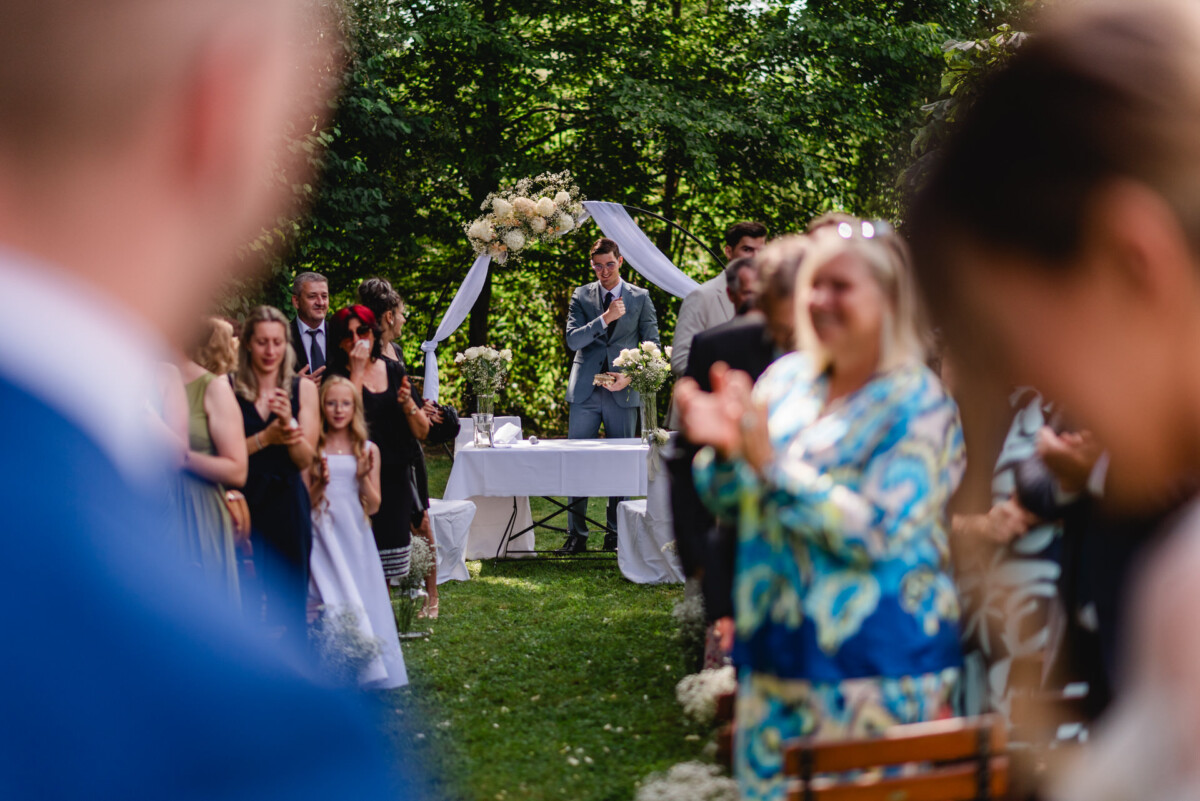 Eine Person in einem grauen Anzug steht an einem Trauungsaltar im Freien und spricht. Gäste, von denen einige unscharf im Vordergrund zu sehen sind, sind versammelt, klatschen und schauen zu.