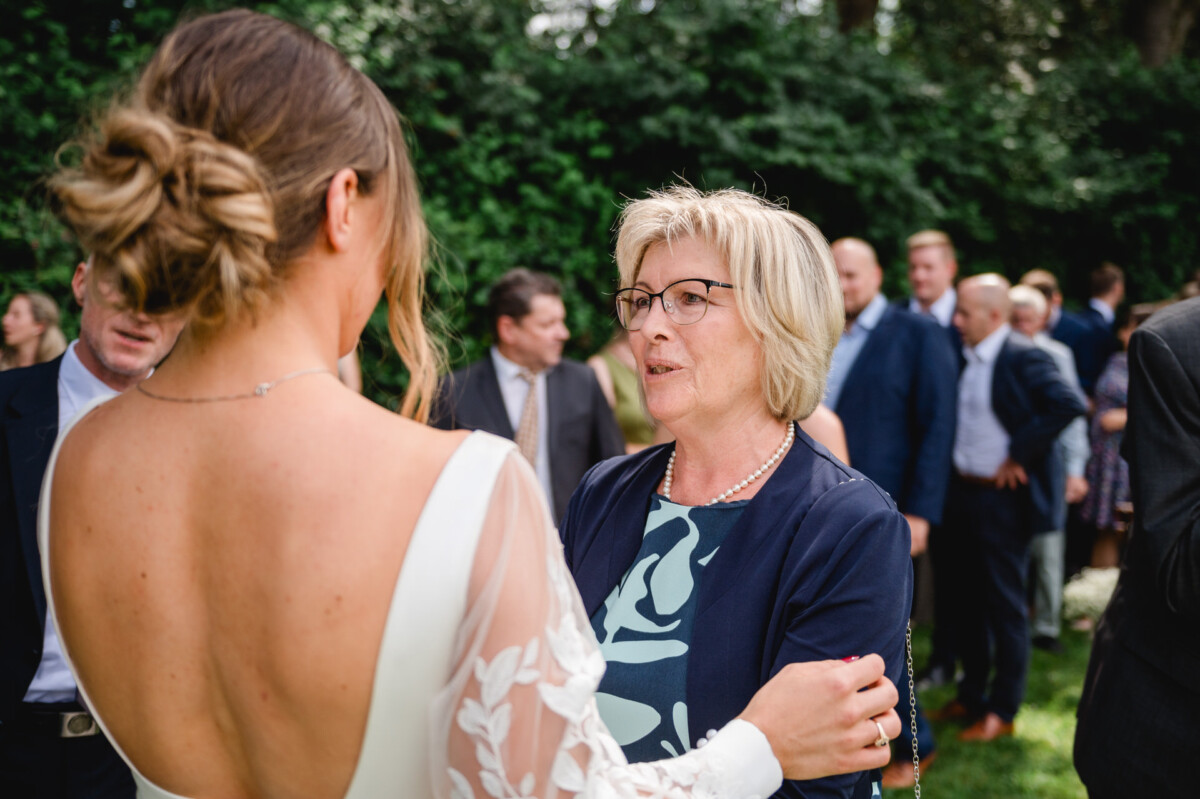 Eine Braut in einem weißen Kleid unterhält sich bei einer Veranstaltung im Freien mit einer älteren Frau in einer dunklen Jacke. Im Hintergrund sind weitere Teilnehmer zu sehen.