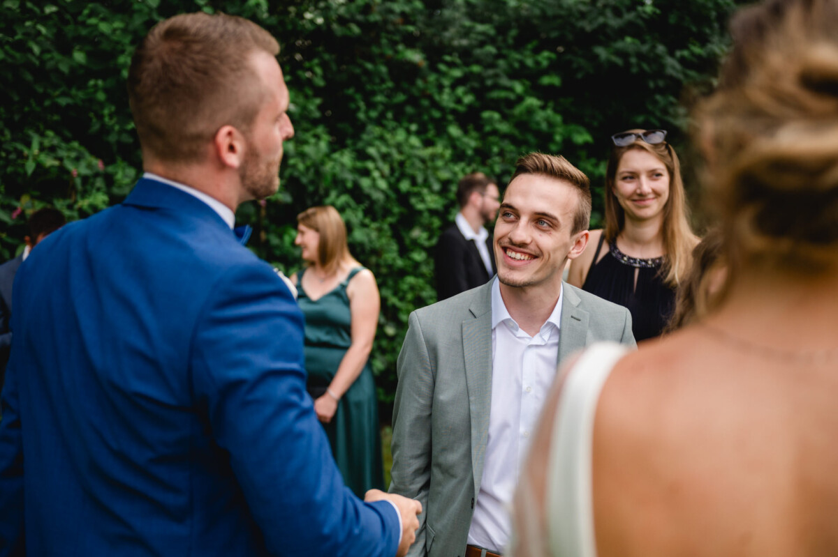 Eine Gruppe von Menschen in formeller Kleidung unterhält sich im Freien; ein Mann im blauen Anzug schüttelt einem lächelnden Mann im grauen Anzug die Hand.