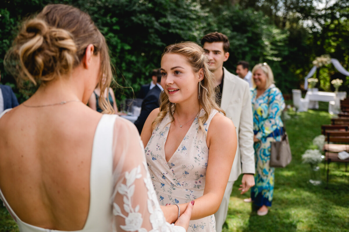 Zwei Frauen halten sich bei einem Treffen im Freien an den Händen und lächeln. Andere Teilnehmer stehen im Hintergrund auf einer Wiese.