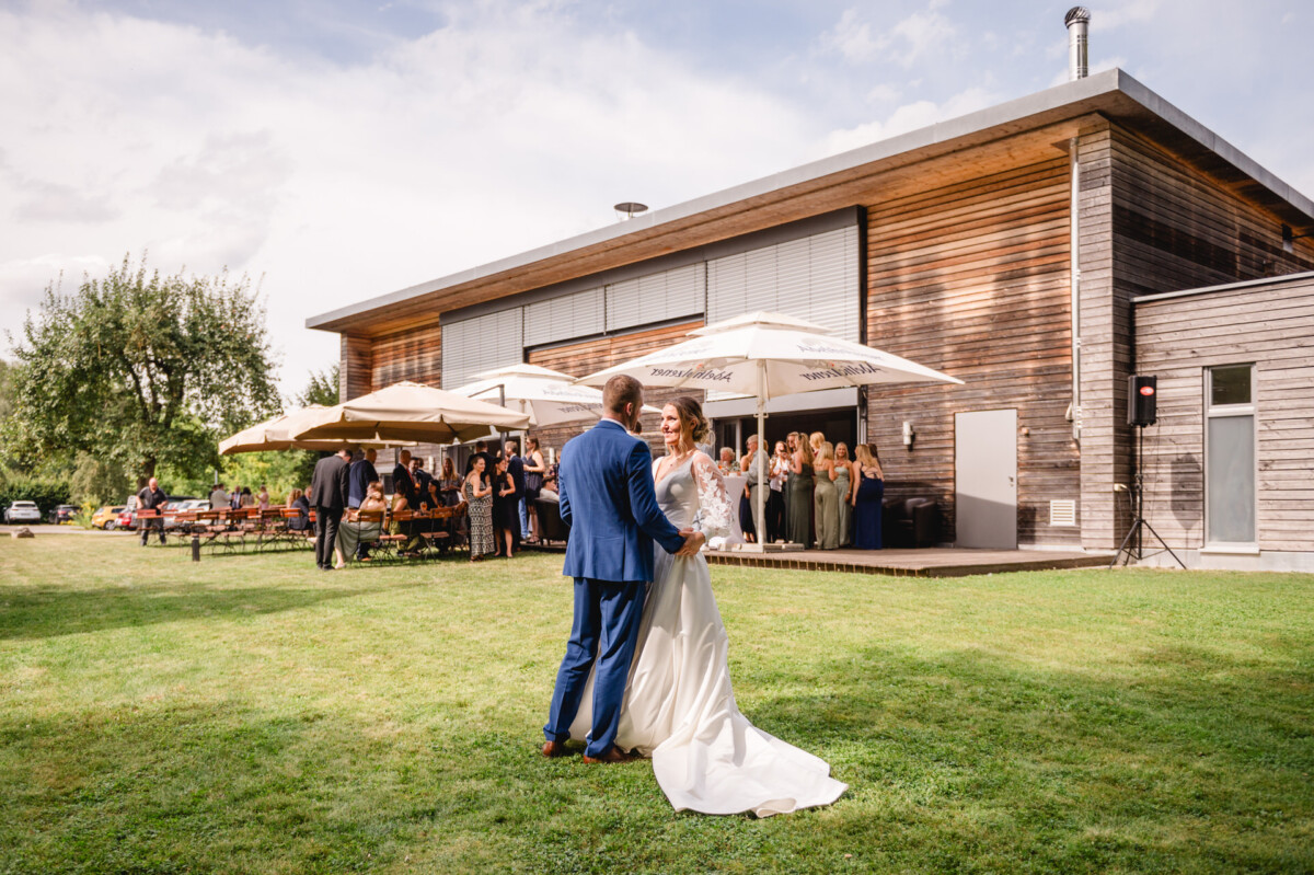 Ein Paar tanzt auf dem Rasen vor einem modernen Holzgebäude, umgeben von Gästen und großen Sonnenschirmen.