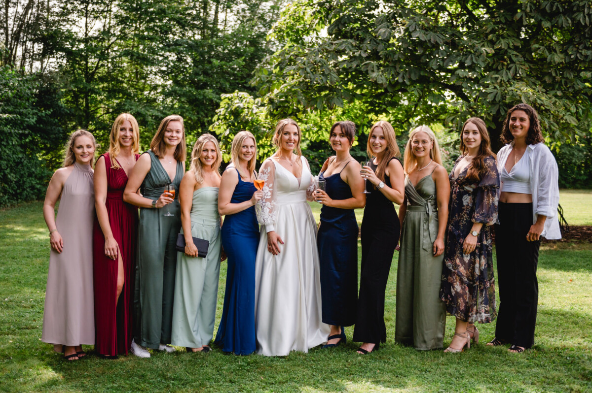 Eine Gruppe von Menschen in Abendgarderobe posiert im Freien auf einer Wiese, im Hintergrund sind Bäume zu sehen.