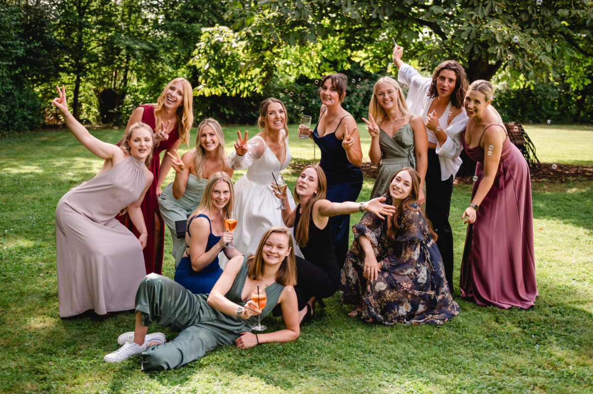 Eine Gruppe von Frauen posiert auf dem Gras. Einige halten Getränke, machen Peace-Zeichen und lächeln. Im Hintergrund sind Bäume und Grünflächen zu sehen.