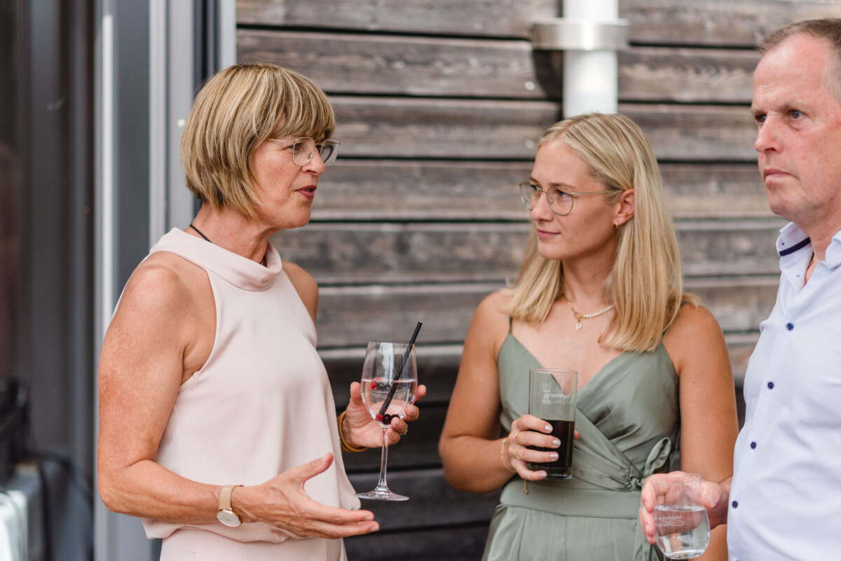 2024.08.09_Martina_Patrick_Hochzeit_Guenzburg_Gundelfingen_Neuhof_am_See_00805 Drei Leute stehen draußen und unterhalten sich. Sie halten Getränke in den Händen. Eine Frau in einem weißen Kleid spricht mit einer Frau in einem grünen Kleid und einem Mann in einem blauen Hemd. Im Hintergrund ist eine Holzverkleidung zu sehen.