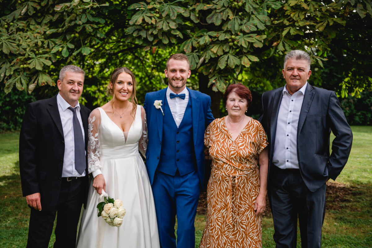 Braut und Bräutigam stehen mit drei älteren Erwachsenen in Abendgarderobe im Freien vor üppigem Grün. Die Braut hält einen Blumenstrauß.