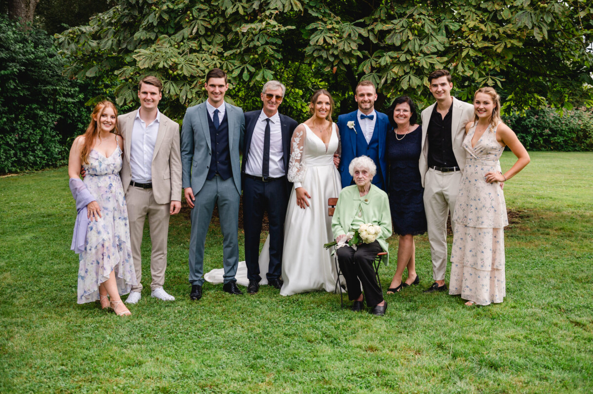 Ein Gruppenporträt einer Hochzeit mit neun stehenden Erwachsenen, zwei in formeller Hochzeitskleidung und einem sitzenden älteren Erwachsenen mit einem Blumenstrauß in der Hand, alle posieren vor einem Baum im Gras.