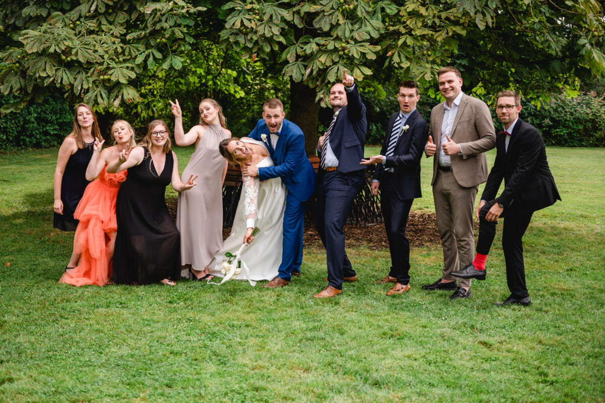Eine Gruppe von Menschen posiert verspielt in Abendgarderobe auf einer Rasenfläche mit Bäumen im Hintergrund.