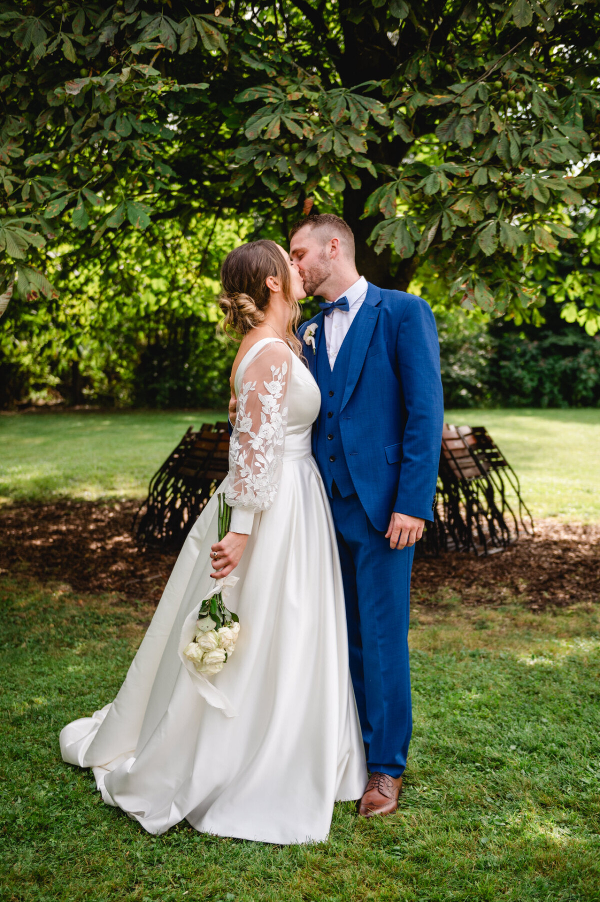 Braut und Bräutigam küssen sich unter einem Baum. Die Braut hält einen Blumenstrauß und trägt ein weißes Kleid, der Bräutigam einen blauen Anzug.