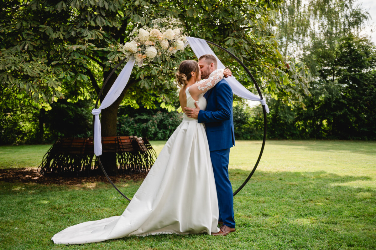 Braut und Bräutigam küssen sich vor einem dekorativen Rundbogen mit weißem Stoff und Blumen in einer Gartenkulisse.