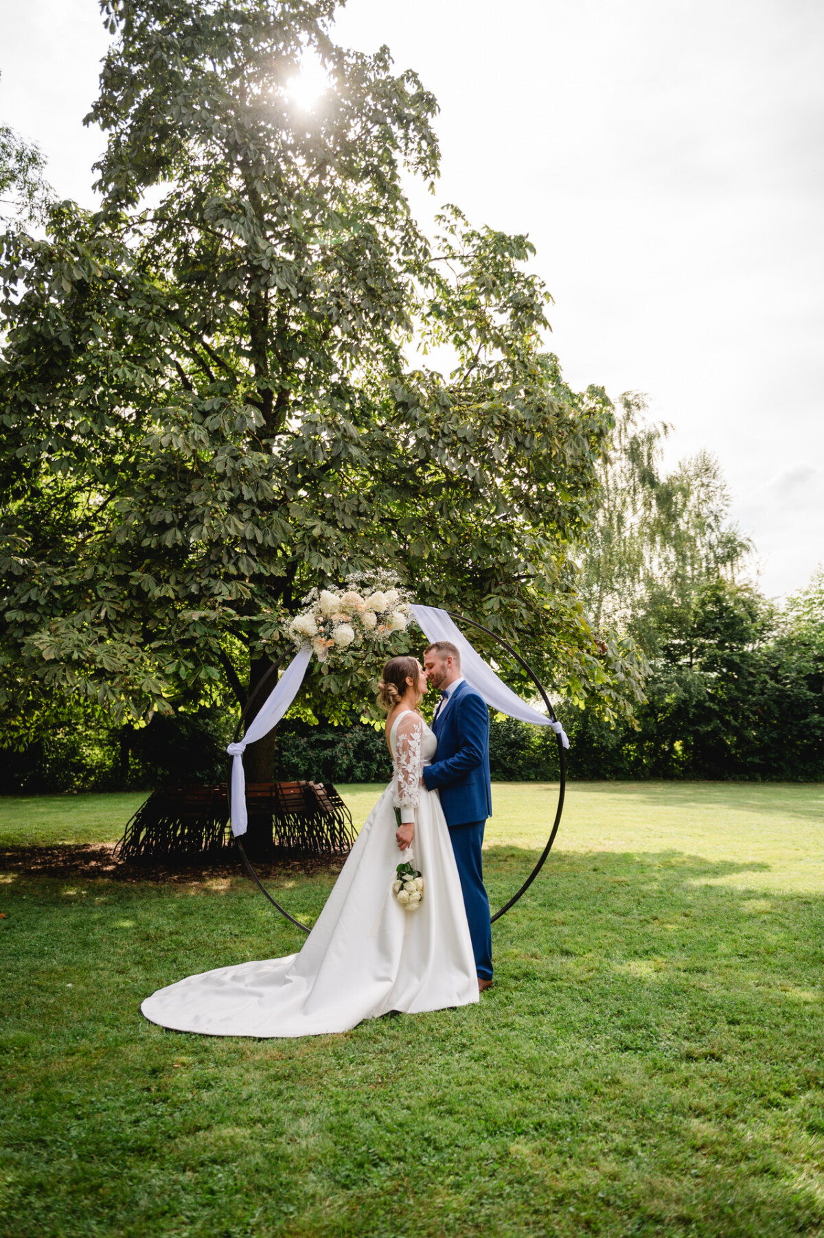 Braut und Bräutigam stehen in einem Garten unter einem großen Baum neben einem runden Hochzeitsbogen, der mit Blumen und Stoff dekoriert ist.