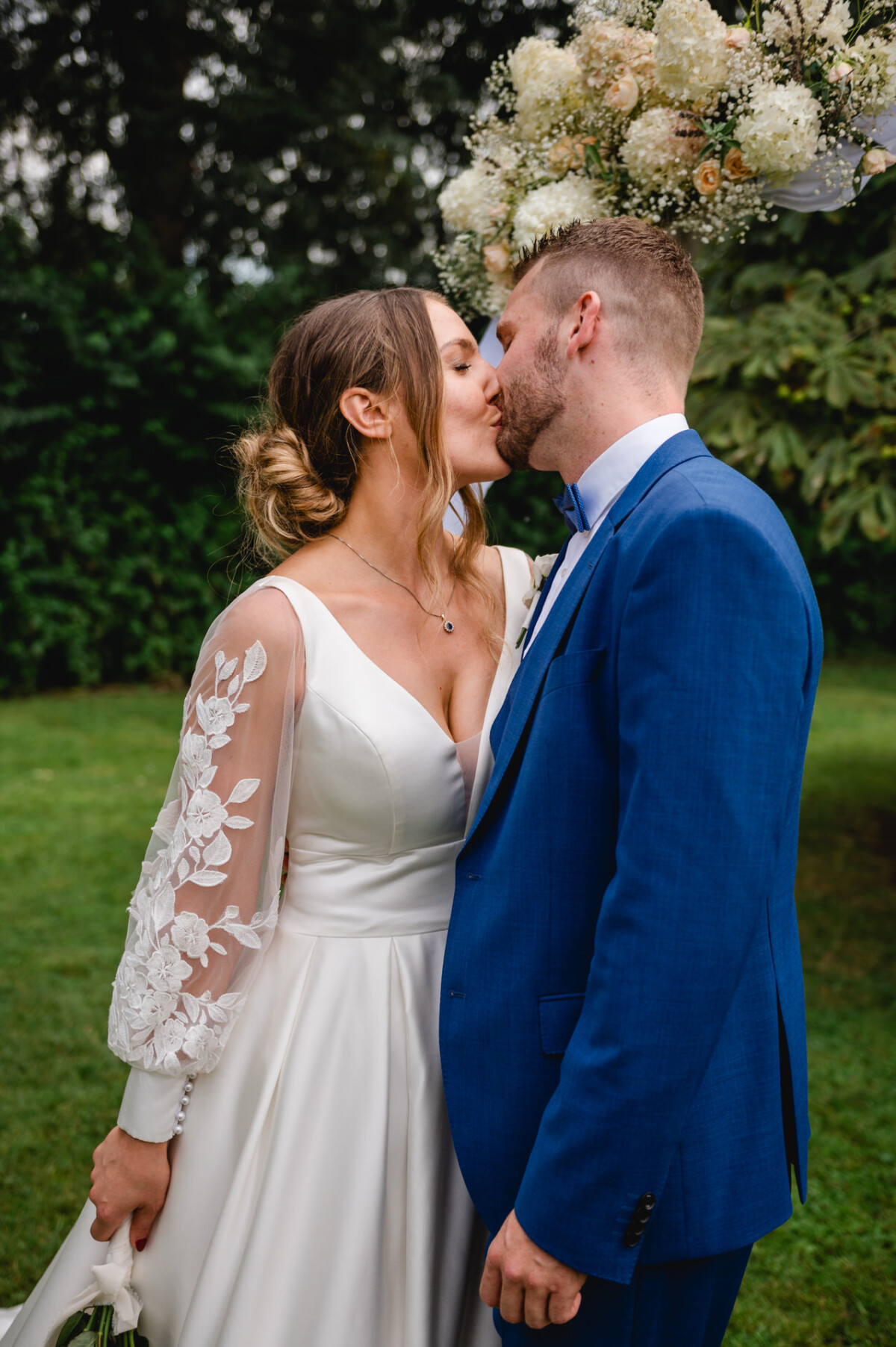 Ein Paar küsst sich im Freien auf einer Wiese. Die Frau trägt ein weißes Kleid mit Spitzenärmeln und der Mann einen blauen Anzug.