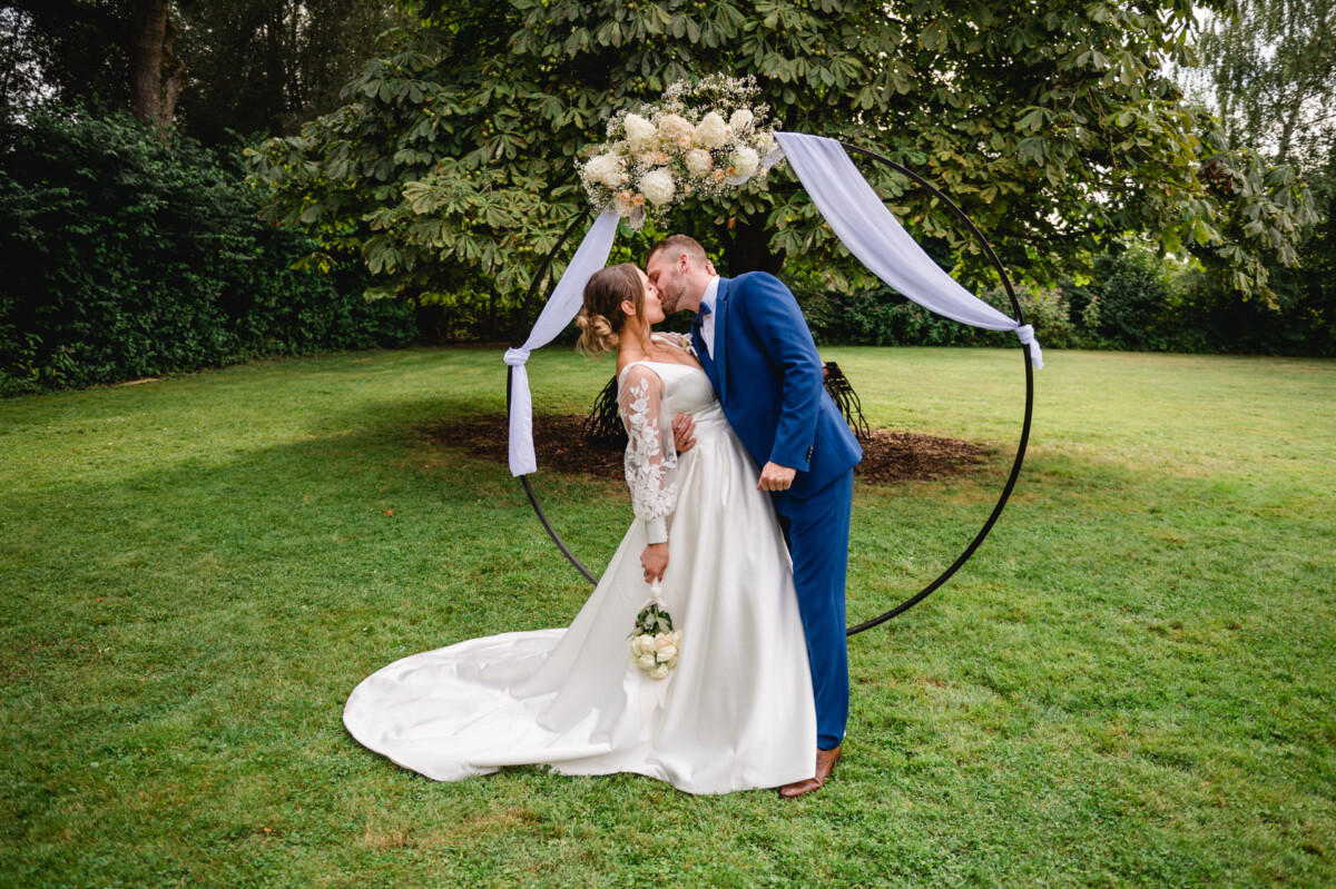 Braut und Bräutigam küssen sich unter einem Blumenbogen in einem Garten. Die Braut trägt ein weißes Kleid und hält einen Blumenstrauß, während der Bräutigam einen blauen Anzug trägt.