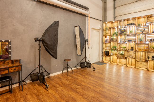 Ein Fotostudio mit zwei Softbox-Leuchten, Holzboden, einem Kosmetikspiegel auf der linken Seite und einer Wandpräsentation mit Kisten mit Pflanzen und Lichtern auf der rechten Seite.