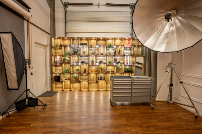 Fotostudio mit Beleuchtungsausrüstung, einer großen Softbox und einem Hintergrund. Holzkisten mit Pflanzen und Dekorationen säumen die Rückwand. Auf dem Holzboden steht ein Metallschrank.