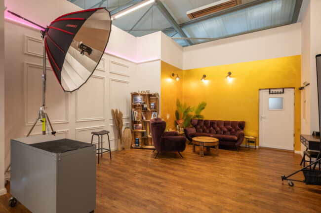 Ein gemütliches Fotostudio mit moderner Einrichtung, bestehend aus großen hellen, braunen Ledersitzen, einem Holztisch, Pflanzen und Wandregalen in einem Raum mit Holzboden und weißen und gelben Wänden.