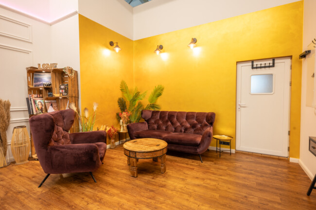 Ein gemütlicher Raum mit einer gelben Wand, braunen Samtsofas, einem runden Couchtisch aus Holz und Topfpflanzen. Im Hintergrund sind Regale mit Dekoartikeln und eine Tür zu sehen. Überall Holzfußboden.