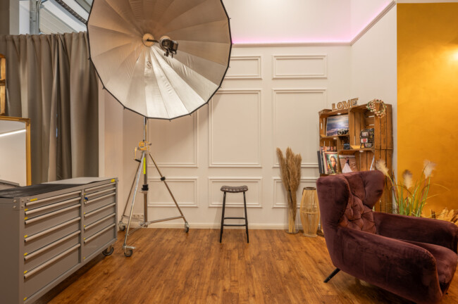 Fotostudio-Christian-Lorenz-Fotografie-Bobingen-00006 Fotostudio-Setup mit großem Schirmlicht, Hocker und Sessel. Zur Dekoration gehören ein Fotodisplay, ein Beistelltisch und Pampasgras vor getäfelten und gelben Wänden.