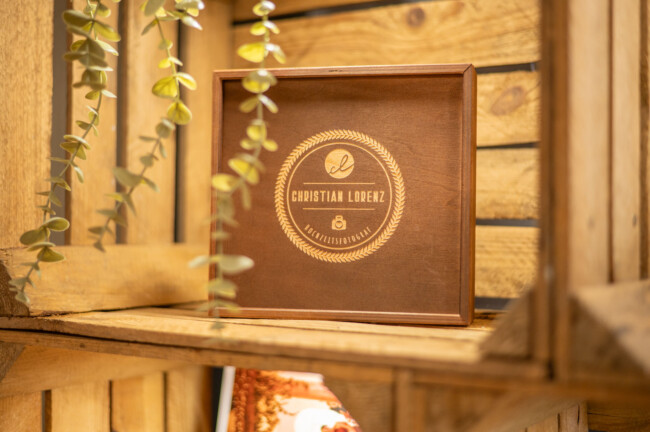Auf einem Regal steht eine Holzkiste mit einem runden Wappen und der Gravur „Christian Lorenz“. Daneben hängen dekorative Blätter.