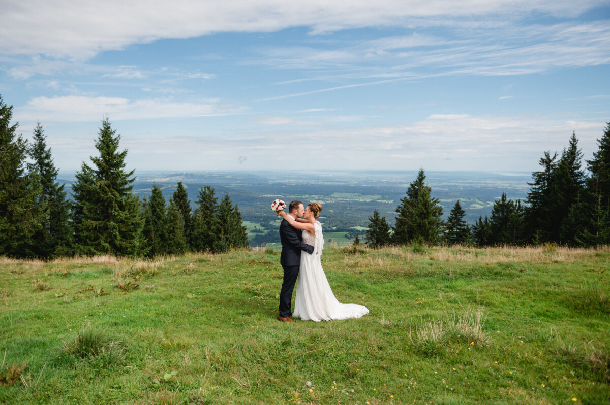 Ein Paar umarmt sich auf einer grasbewachsenen Hügelkuppe mit malerischer Aussicht auf Bäume und Felder unter einem teilweise bewölkten Himmel.