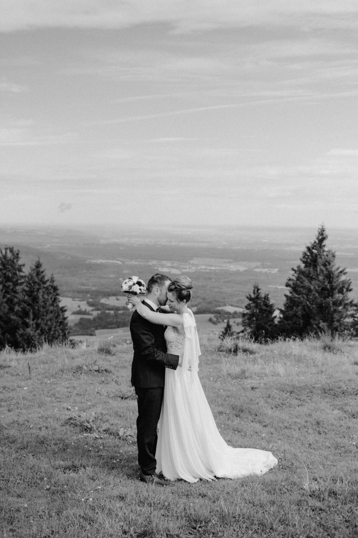 Braut und Bräutigam umarmen sich auf einem grasbewachsenen Hügel, vor der Kulisse von Bäumen und einer fernen Landschaft unter einem bewölkten Himmel. Das Bild ist in Schwarzweiß.