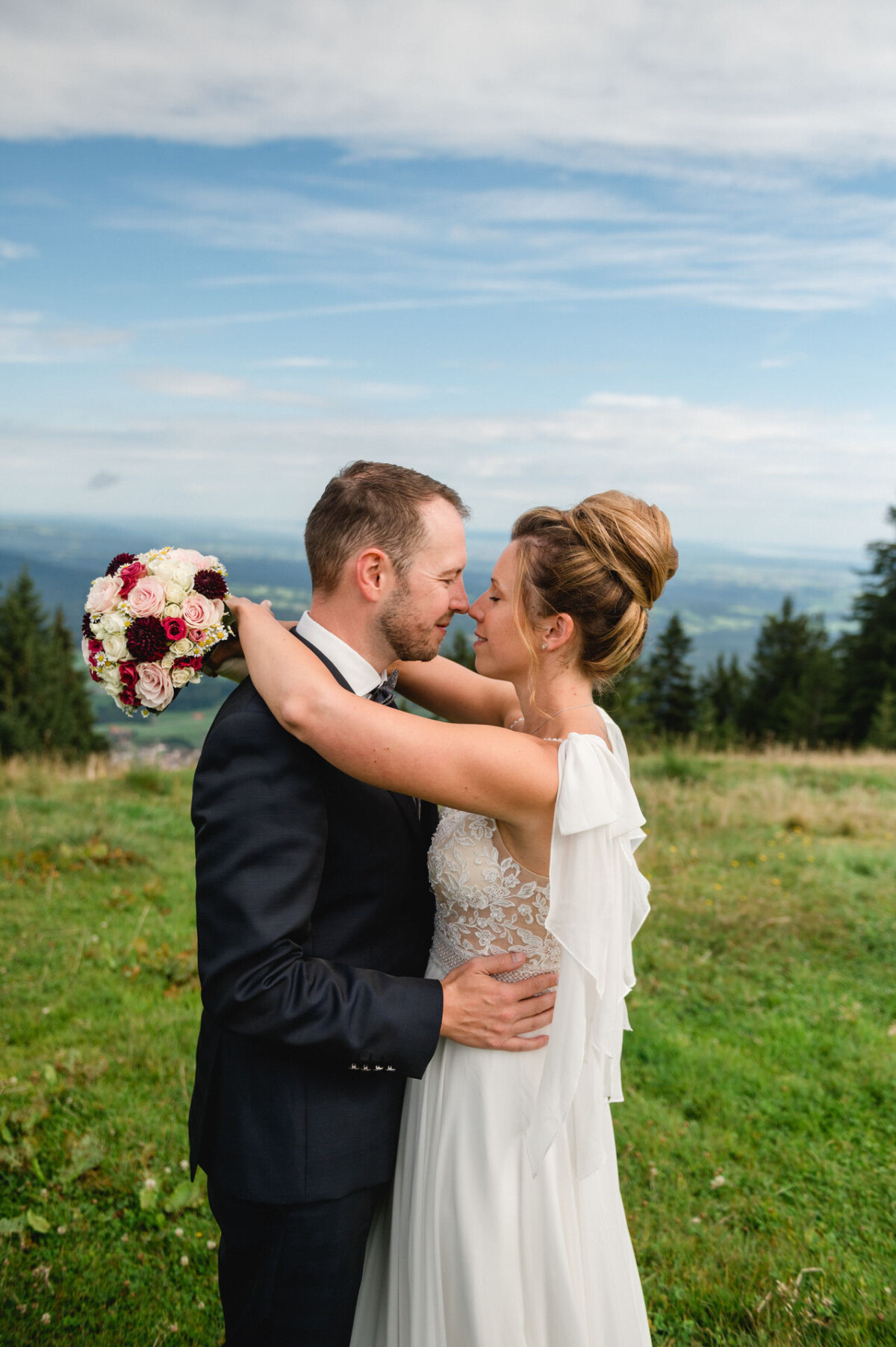 Ein Paar in Hochzeitskleidung steht eng beieinander auf einer Wiese und hält einen Blumenstrauß in den Händen. Der Mann trägt einen Anzug und die Frau ein weißes Kleid. Im Hintergrund sind Bäume und der Himmel zu sehen.