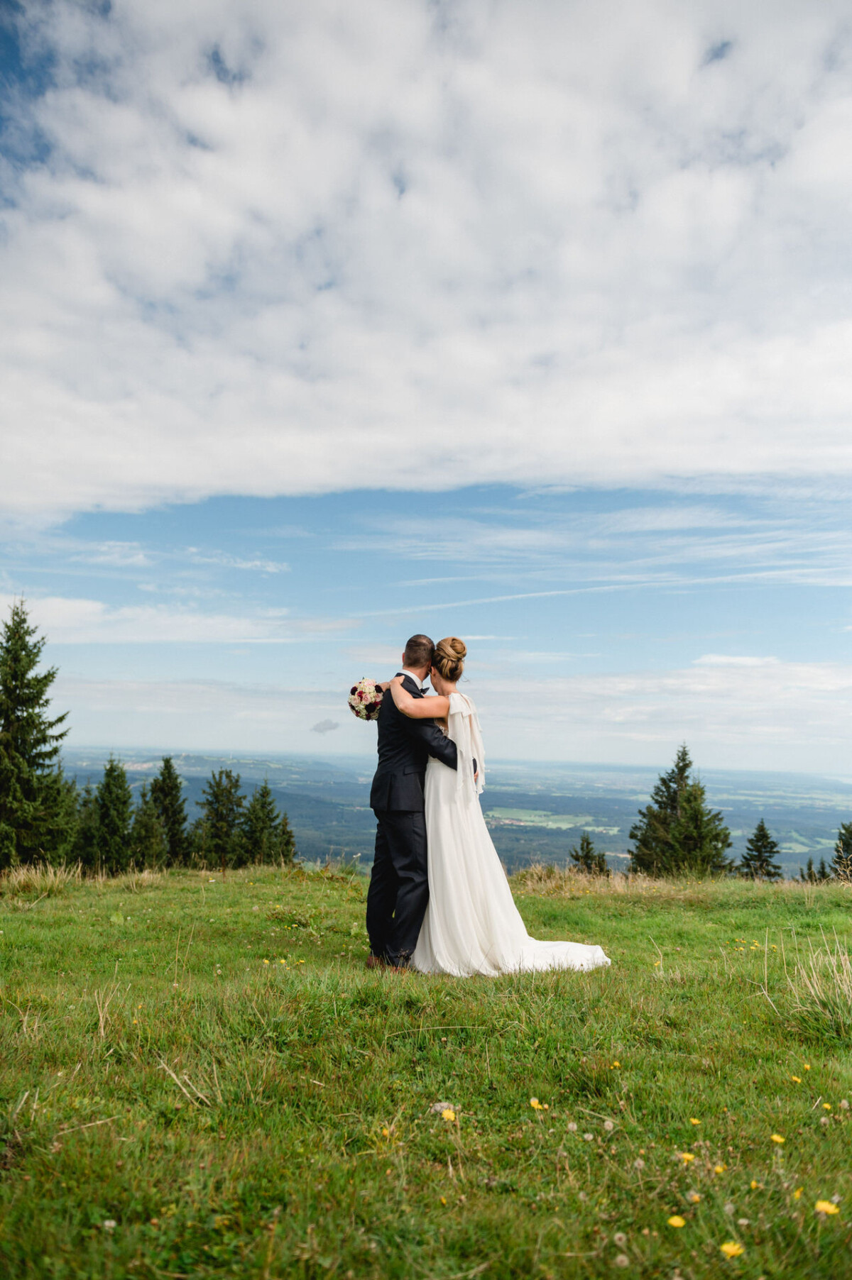 Braut und Bräutigam umarmen sich auf einer grasbewachsenen Hügelkuppe, im Hintergrund eine malerische Landschaft und ein bewölkter Himmel.
