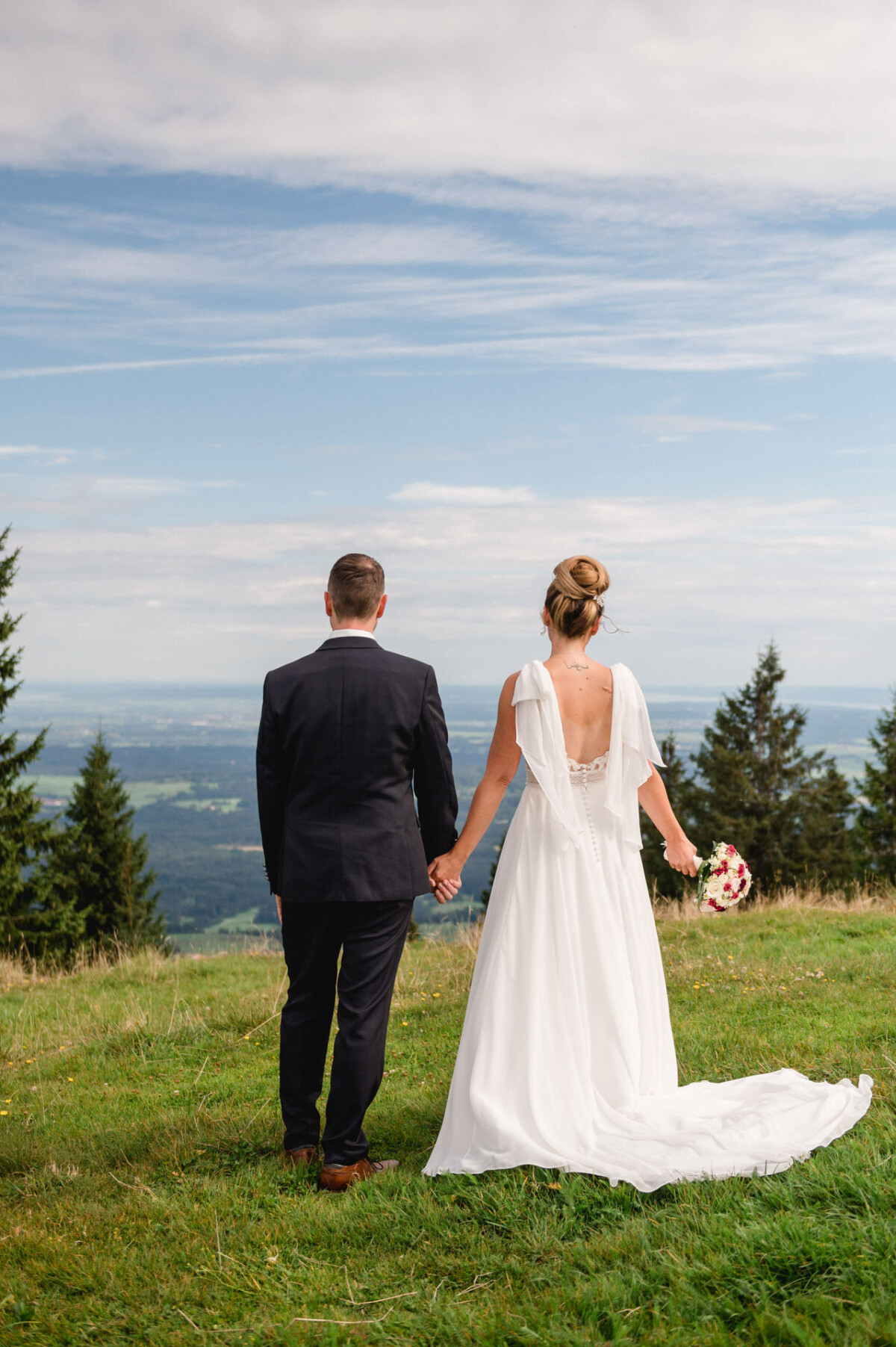 Braut und Bräutigam stehen Händchen haltend und mit abgewandtem Blick auf einem grasbewachsenen Hügel und blicken auf eine Landschaft mit Bäumen und einem fernen Horizont unter einem teilweise bewölkten Himmel.