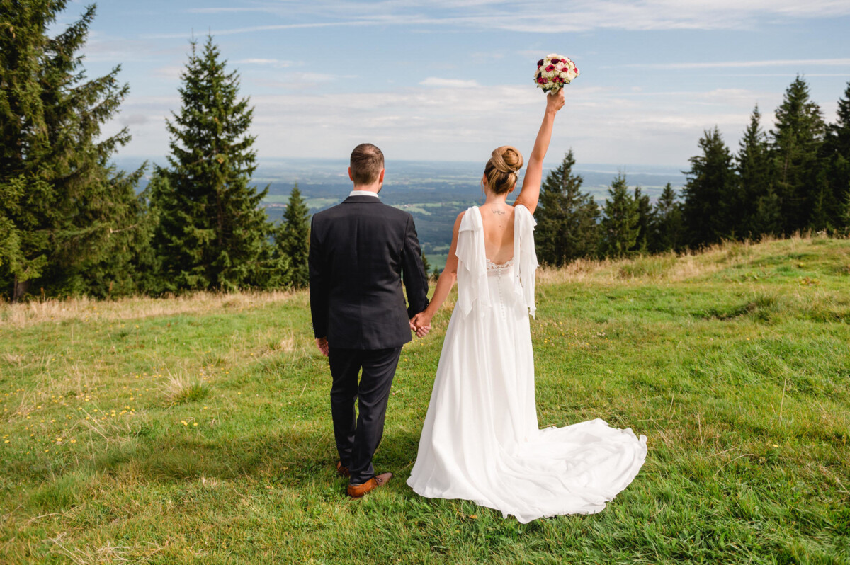 Braut und Bräutigam stehen auf einem grasbewachsenen Hügel und halten sich an den Händen. Die Braut hält einen Blumenstrauß hoch. Im Hintergrund sind Kiefern und eine malerische Landschaft zu sehen.