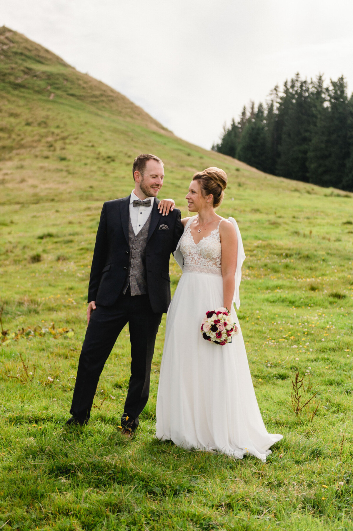 Ein Paar im Hochzeitskleid steht auf einem grasbewachsenen Hügel und lächelt sich an. Die Braut hält einen Blumenstrauß in der Hand, im Hintergrund sind Bäume zu sehen.