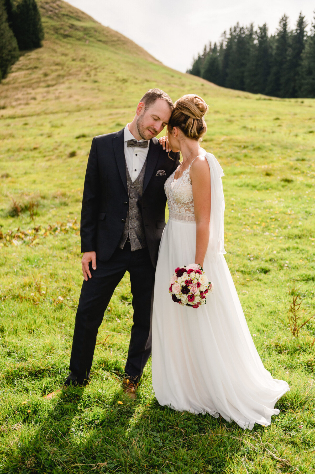 Braut und Bräutigam stehen zusammen auf einem grasbewachsenen Hügel. Sie hält einen Strauß roter und weißer Blumen in der Hand. Beide tragen formelle Hochzeitskleidung.