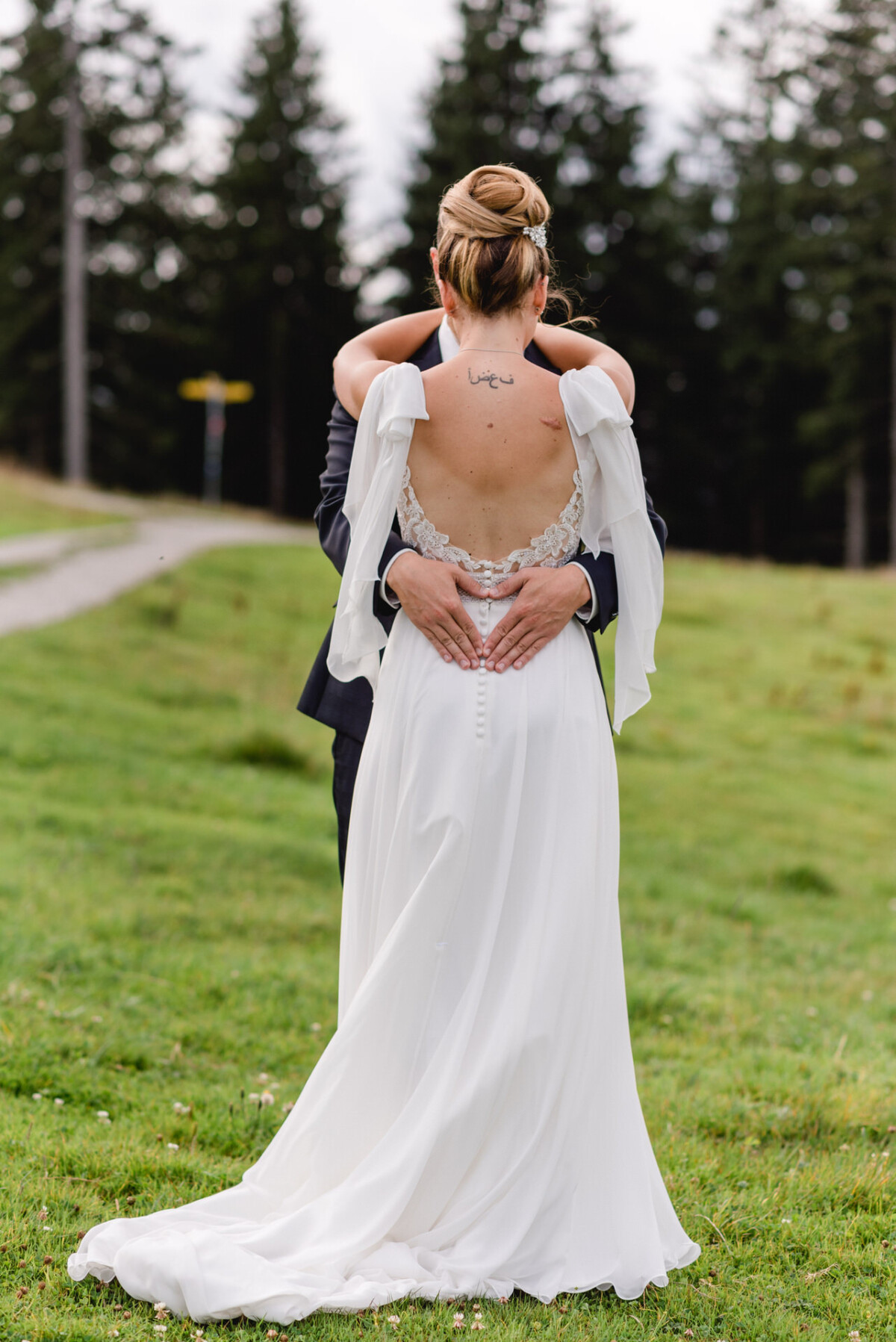 Ein Paar umarmt sich draußen auf einer Wiese, von hinten zu sehen. Die Frau trägt ein weißes Kleid und hat eine Hochsteckfrisur, während der Mann einen dunklen Anzug trägt. Im Hintergrund sind Bäume zu sehen.