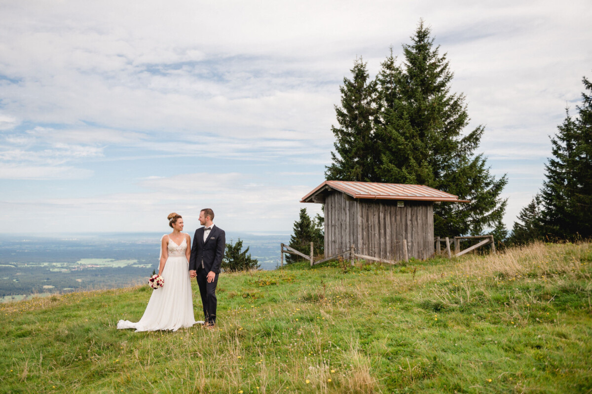 Eine Braut und ein Bräutigam stehen Händchen haltend auf einer Wiese in der Nähe eines kleinen Holzschuppens, vor der Kulisse von Bäumen und einer weiten Landschaft.