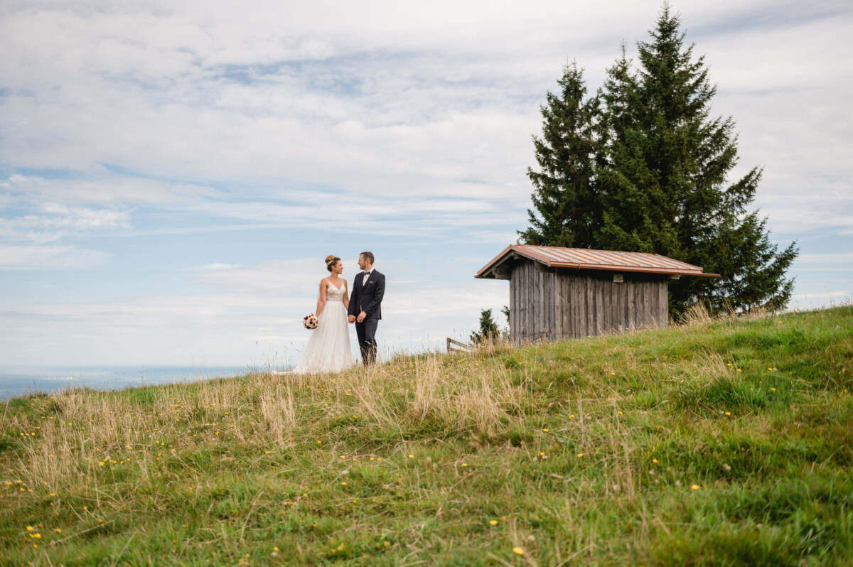Braut und Bräutigam stehen unter einem bewölkten Himmel auf einem grasbewachsenen Hügel in der Nähe eines kleinen Holzschuppens mit Bäumen im Hintergrund.