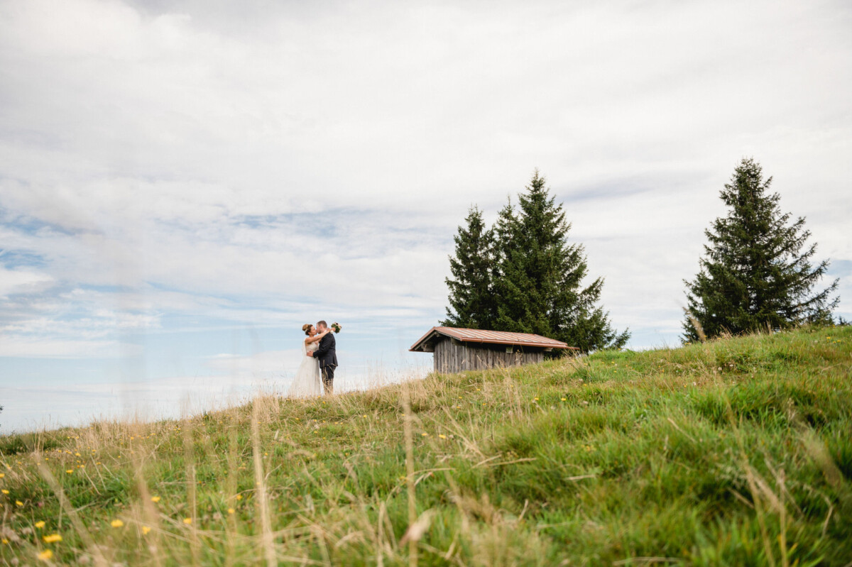 Ein Paar im Hochzeitskleid steht auf einem grasbewachsenen Hügel in der Nähe einer kleinen Holzhütte, umgeben von hohen Bäumen und unter einem bewölkten Himmel.