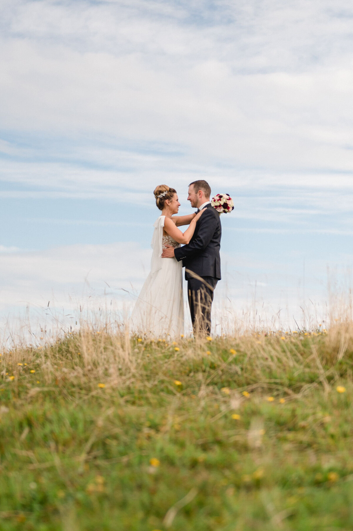 Braut und Bräutigam stehen umarmend auf einem grasbewachsenen Hügel unter einem teilweise bewölkten Himmel. Die Braut hält einen Blumenstrauß.