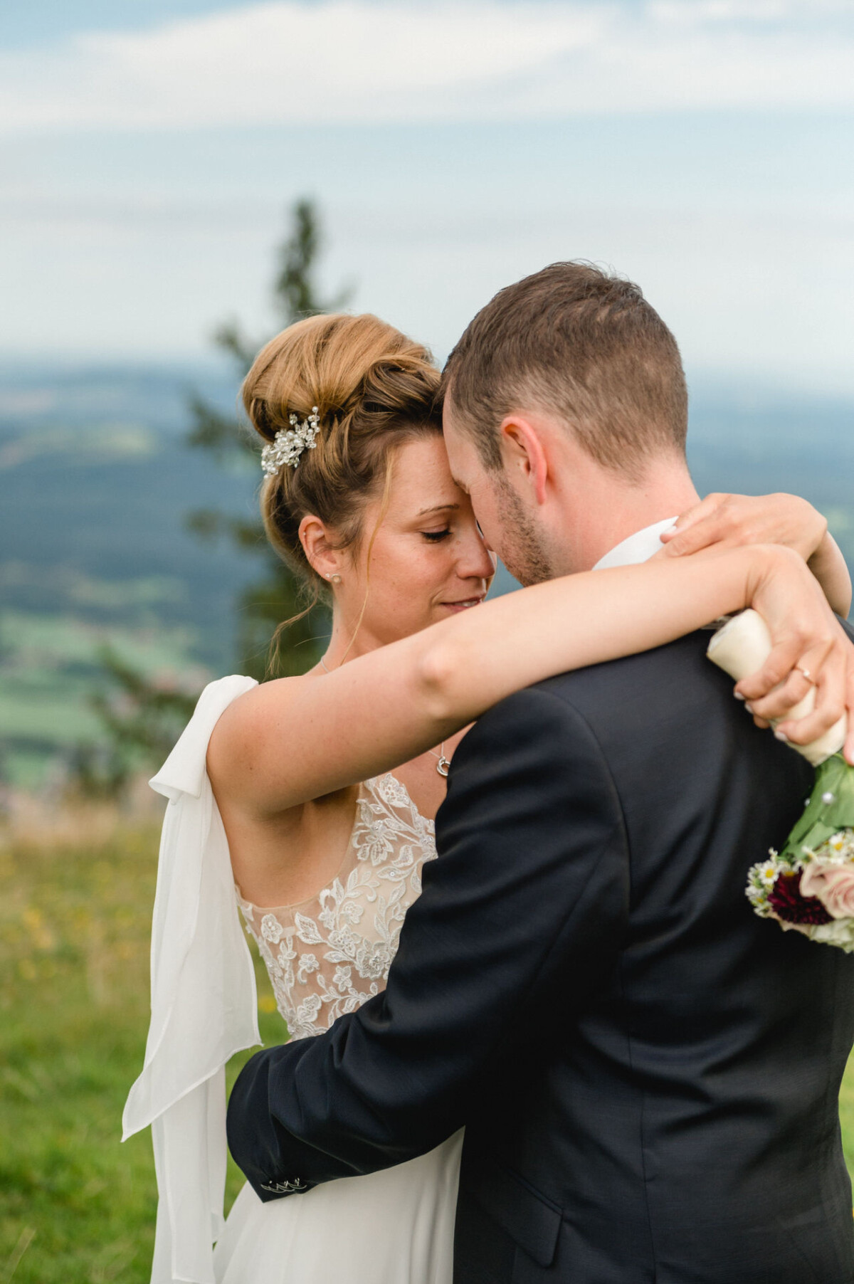 Braut und Bräutigam umarmen sich auf einem grasbewachsenen Hügel mit einer malerischen Landschaft im Hintergrund. Die Braut hält einen Blumenstrauß und beide sind in Abendgarderobe gekleidet.