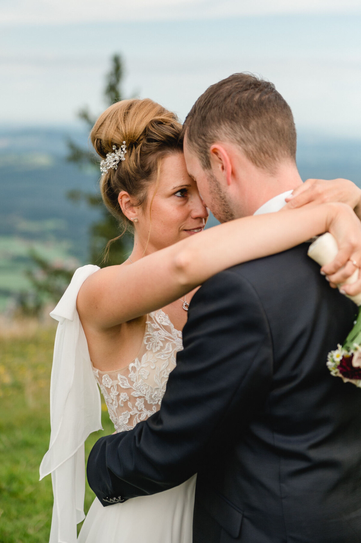 Braut und Bräutigam umarmen sich im Freien und stehen sich dicht gegenüber. Die Braut hält einen Blumenstrauß und beide tragen formelle Hochzeitskleidung mit einer malerischen Landschaft im Hintergrund.