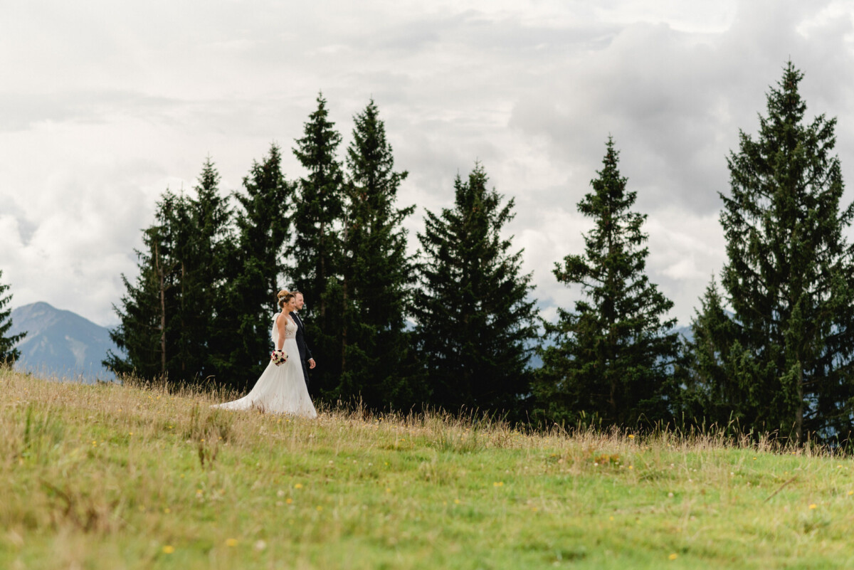 Eine Frau in einem weißen Kleid geht durch eine Wiese mit immergrünen Bäumen im Hintergrund.