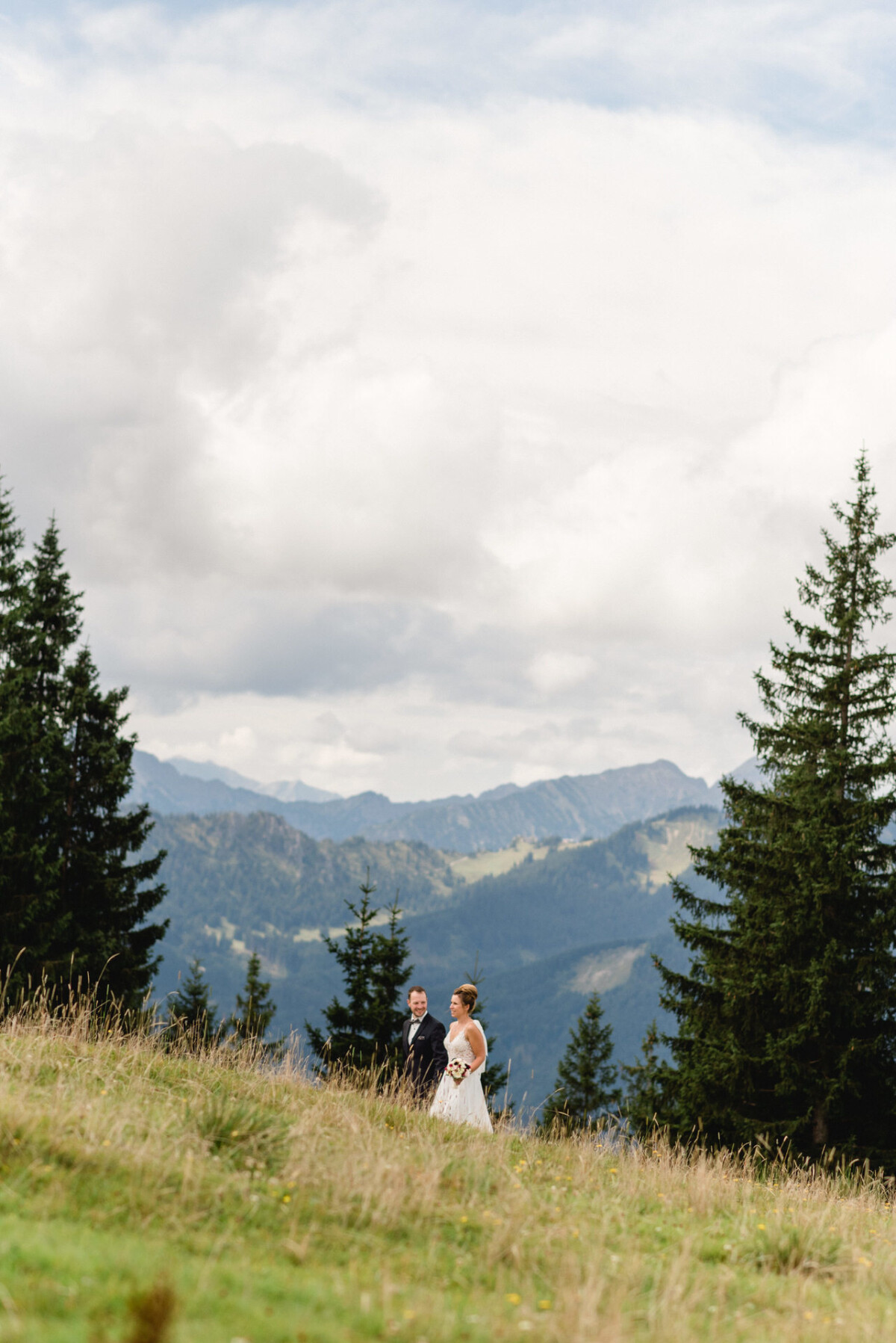Eine Braut und ein Bräutigam gehen unter einem bewölkten Himmel durch eine Wiese mit Bergen und Bäumen im Hintergrund.