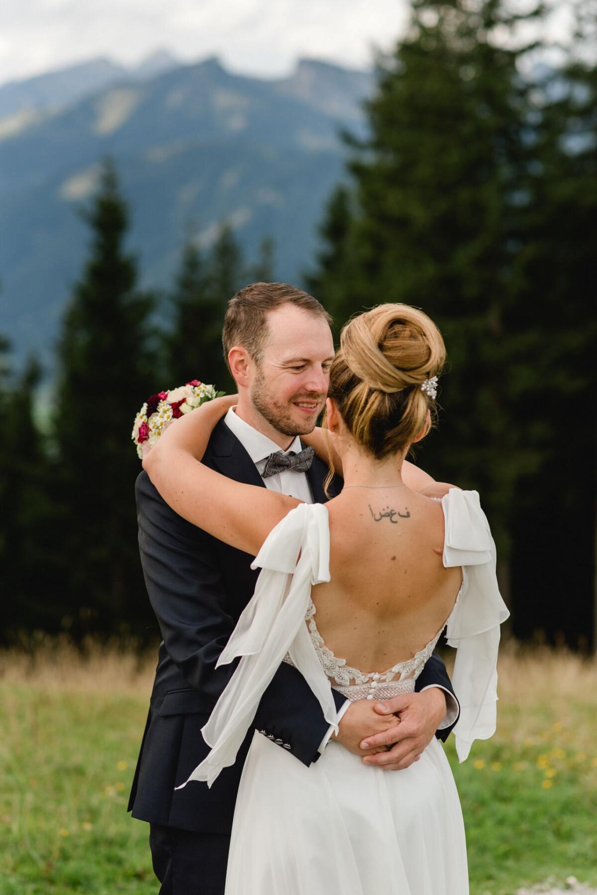 Ein Paar umarmt sich im Freien, mit Bergen und Bäumen im Hintergrund. Die Frau trägt ein weißes Kleid und der Mann einen dunklen Anzug. Sie hält einen Blumenstrauß und hat eine Tätowierung auf dem Rücken.