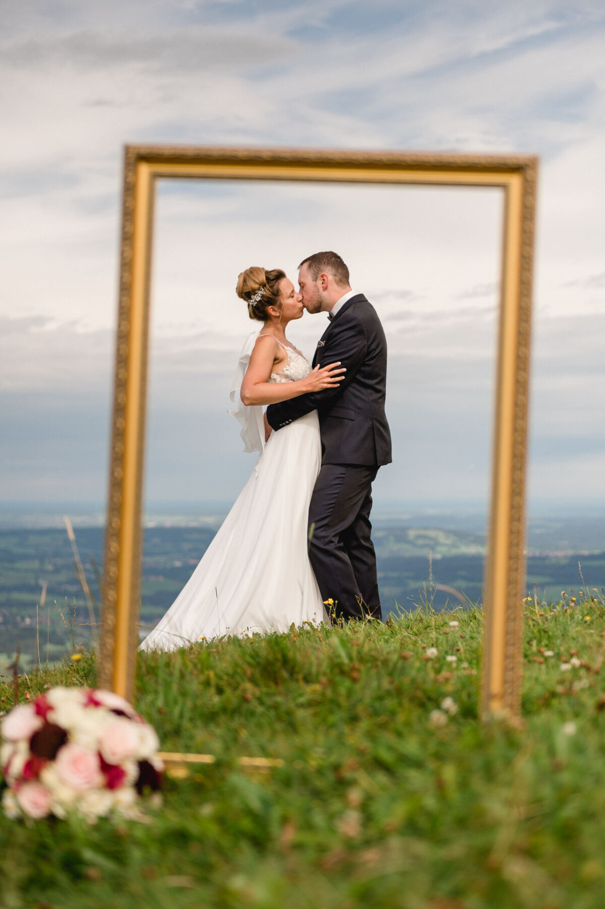 Ein Brautpaar küsst sich auf einer grasbewachsenen Hügelkuppe, eingerahmt von einem leeren Bilderrahmen.
