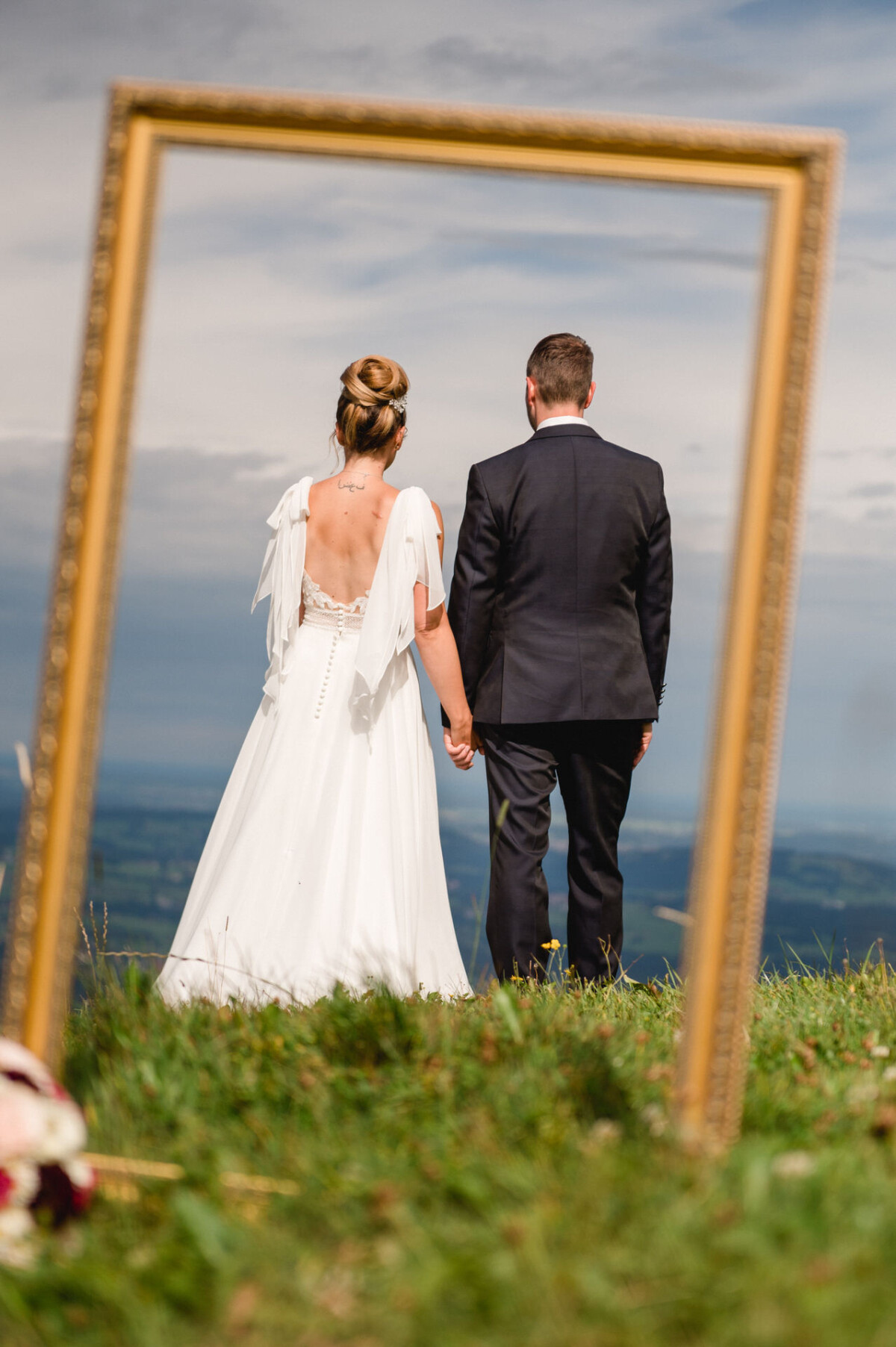 Eine Braut und ein Bräutigam im Hochzeitskleid stehen Hand in Hand auf einer grasbewachsenen Hügelkuppe, eingerahmt von einem leeren Bilderrahmen, mit einem bewölkten Himmel und einer Landschaft im Hintergrund.
