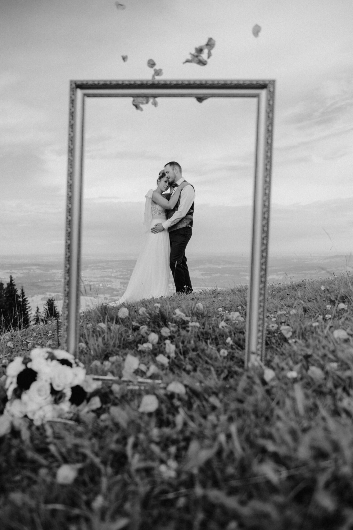 Ein Paar umarmt sich auf einem grasbewachsenen Hügel, reflektiert in einem großen Standspiegel. In der Nähe sind Rosen verstreut und Blütenblätter schweben in der Luft. Die Szene ist in Schwarzweiß festgehalten.