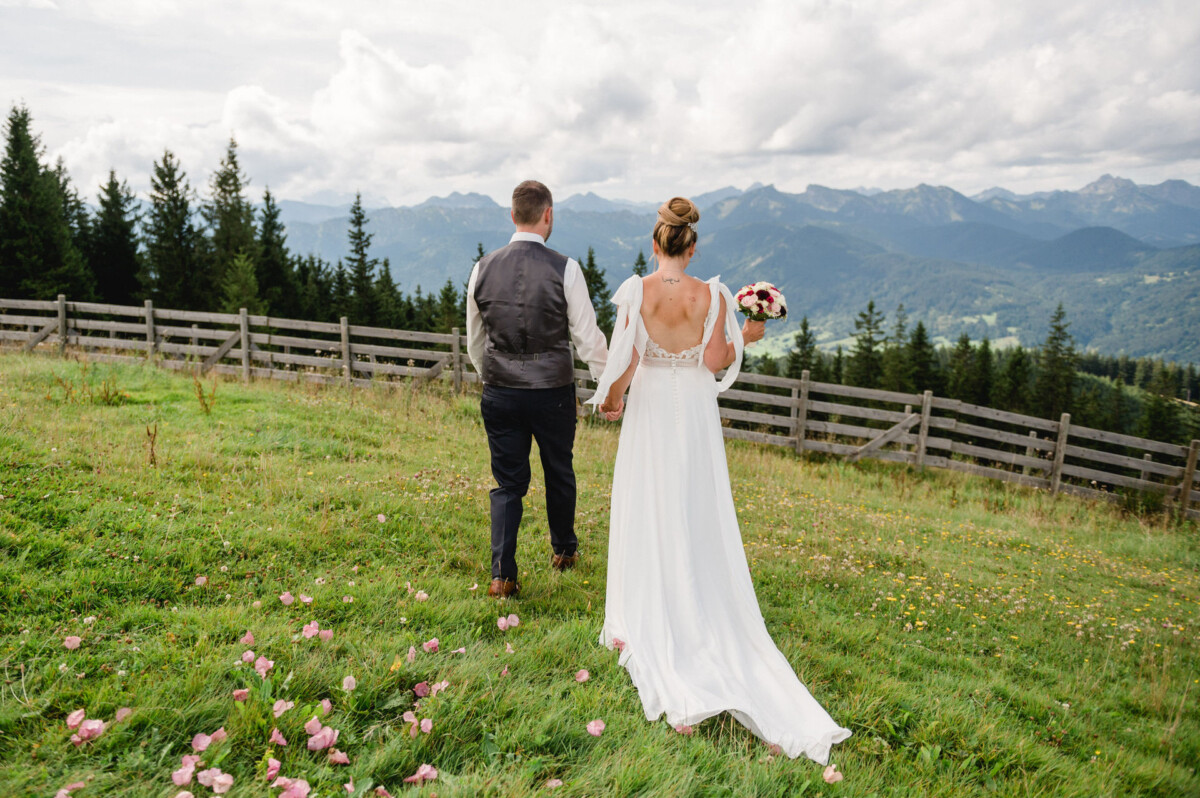 Eine Braut und ein Bräutigam gehen Hand in Hand über eine Wiese mit Bergen im Hintergrund und verstreuten rosa Blumen auf dem Boden.