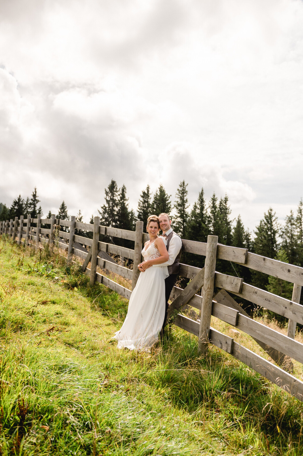 Ein Paar in Hochzeitskleidung steht neben einem Holzzaun auf einer Wiese mit hohen Bäumen und einem bewölkten Himmel im Hintergrund.